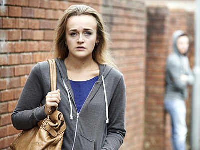 Ein Mädchen mit blonden Haaren, das besorgt aussieht, steht vor einer Ziegelmauer. Im Hintergrund ist eine unscharfe Person zu sehen.